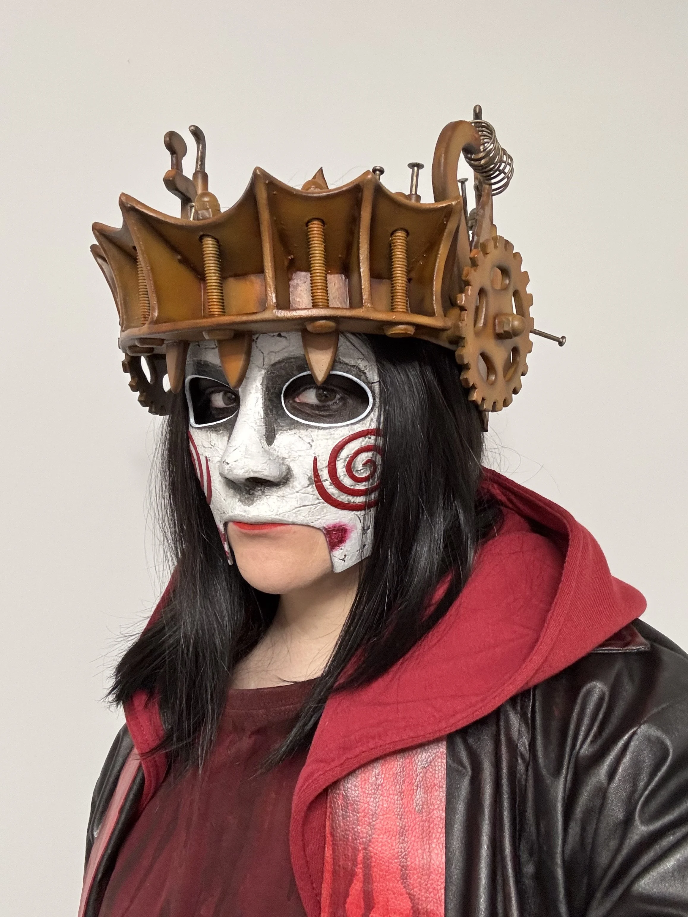 Person wearing a white face mask with red spiral designs, a steampunk-inspired crown with gears and springs, dark hair, a red hoodie, and a black leather jacket.