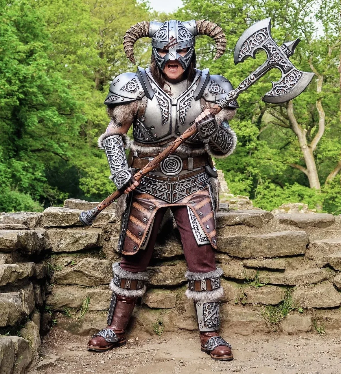 Person dressed as a warrior in armor with horned helmet, holding a large decorated axe, standing outdoors on stone ruins, surrounded by green trees.