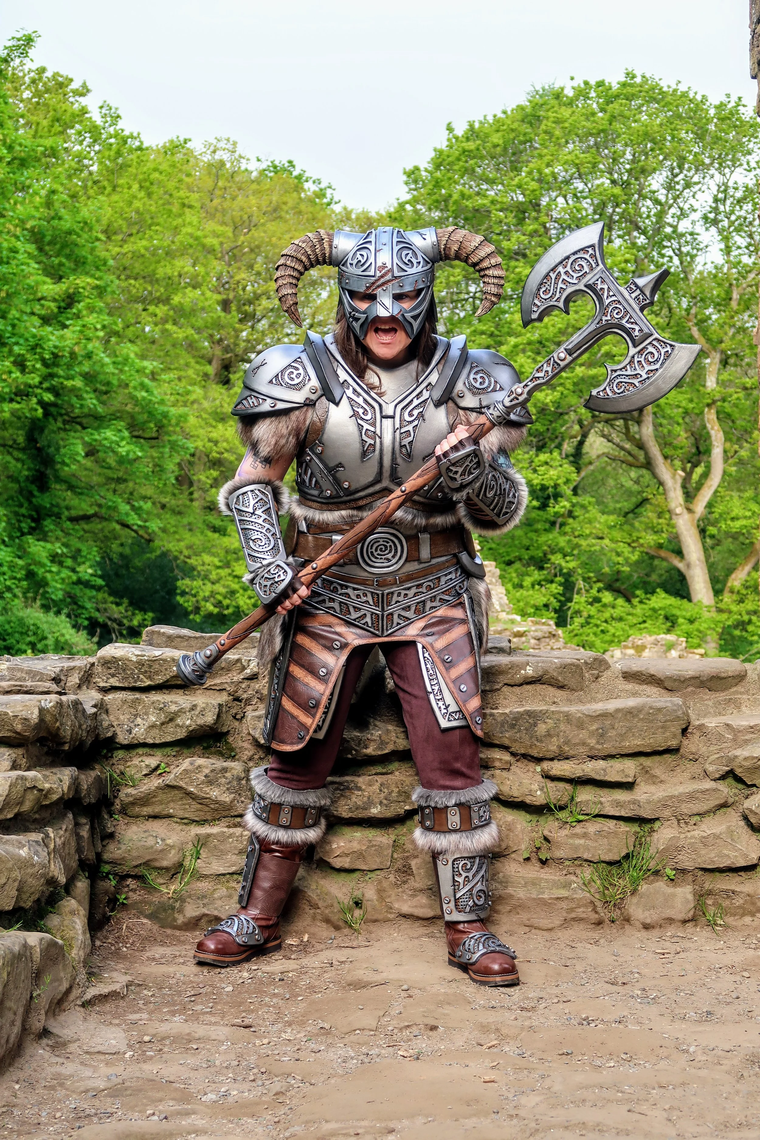 A person dressed as a fantasy warrior in ornate armor and helmet with horns, holding a detailed axe, standing on stone steps outdoors surrounded by green trees.