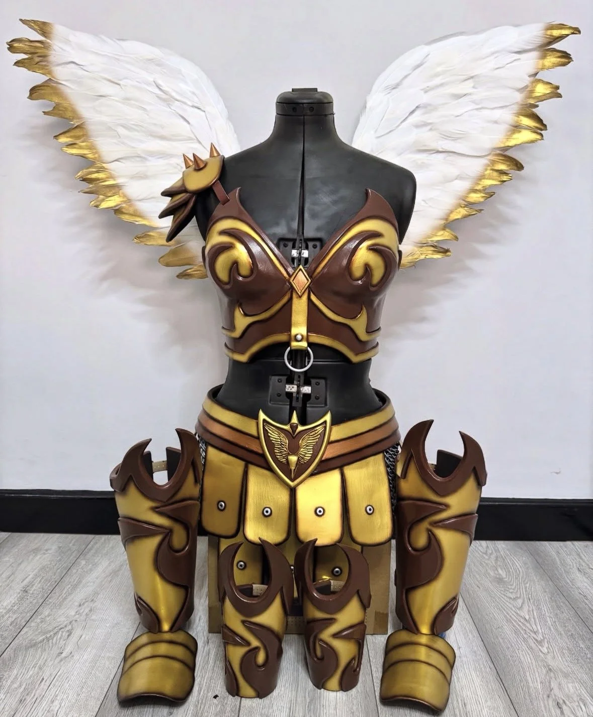A costume display featuring a fantasy warrior armor with large feathered wings attached to the back. The armor is primarily yellow and brown with ornate patterns, a winged emblem on the chest, and detailed arm and leg guards. The wings are white with