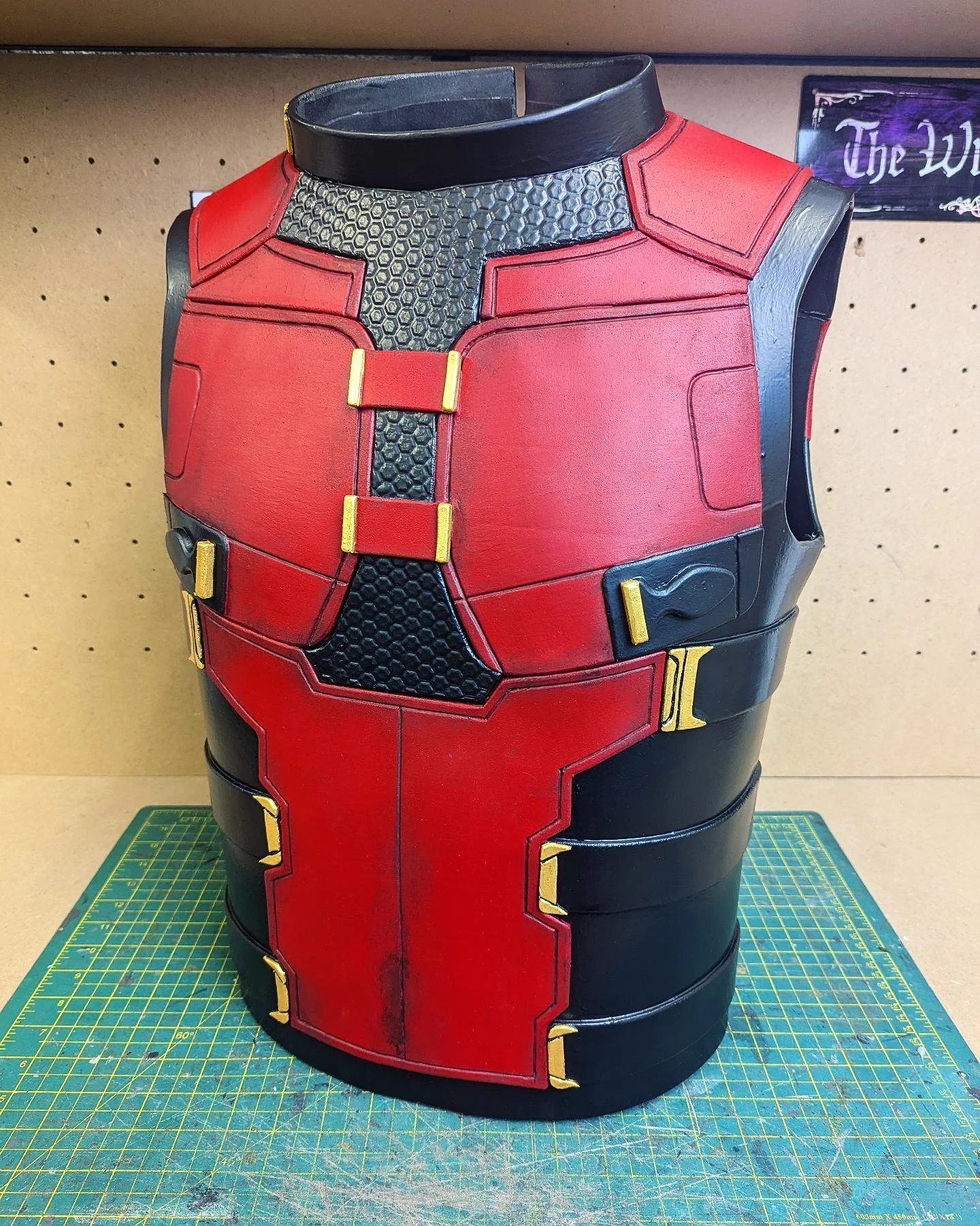 A detailed costume armor piece resembling Deadpool's suit, with black, red, and gold accents, displayed on a green cutting mat on a workbench. The armor has a textured honeycomb pattern on the upper chest area, gold buckles, and is segmented with various panels.