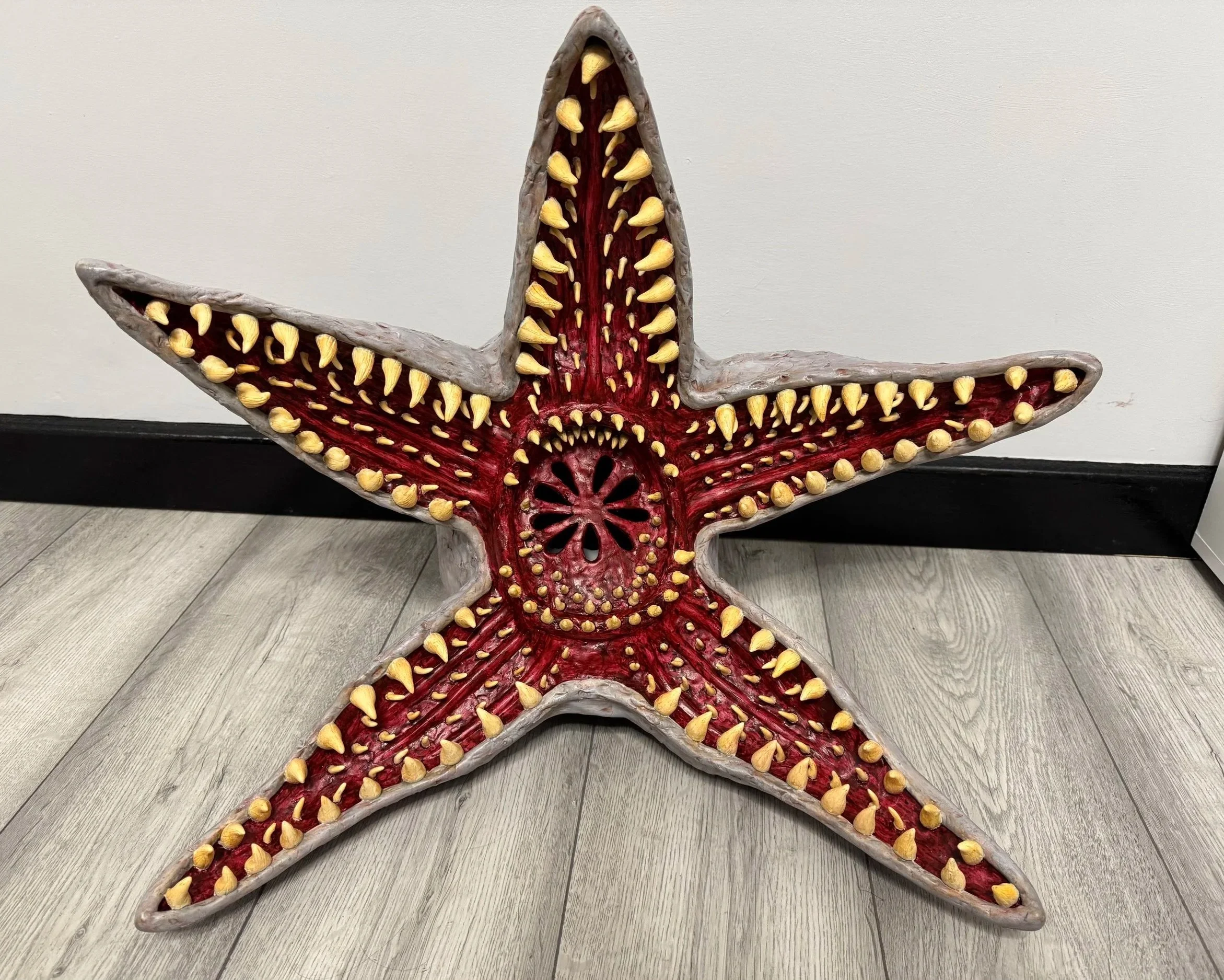 A starfish-shaped sculpture with a red interior, gray outer edges, and yellowish protruding cone-shaped elements inside, placed against a plain wall on a wooden floor.