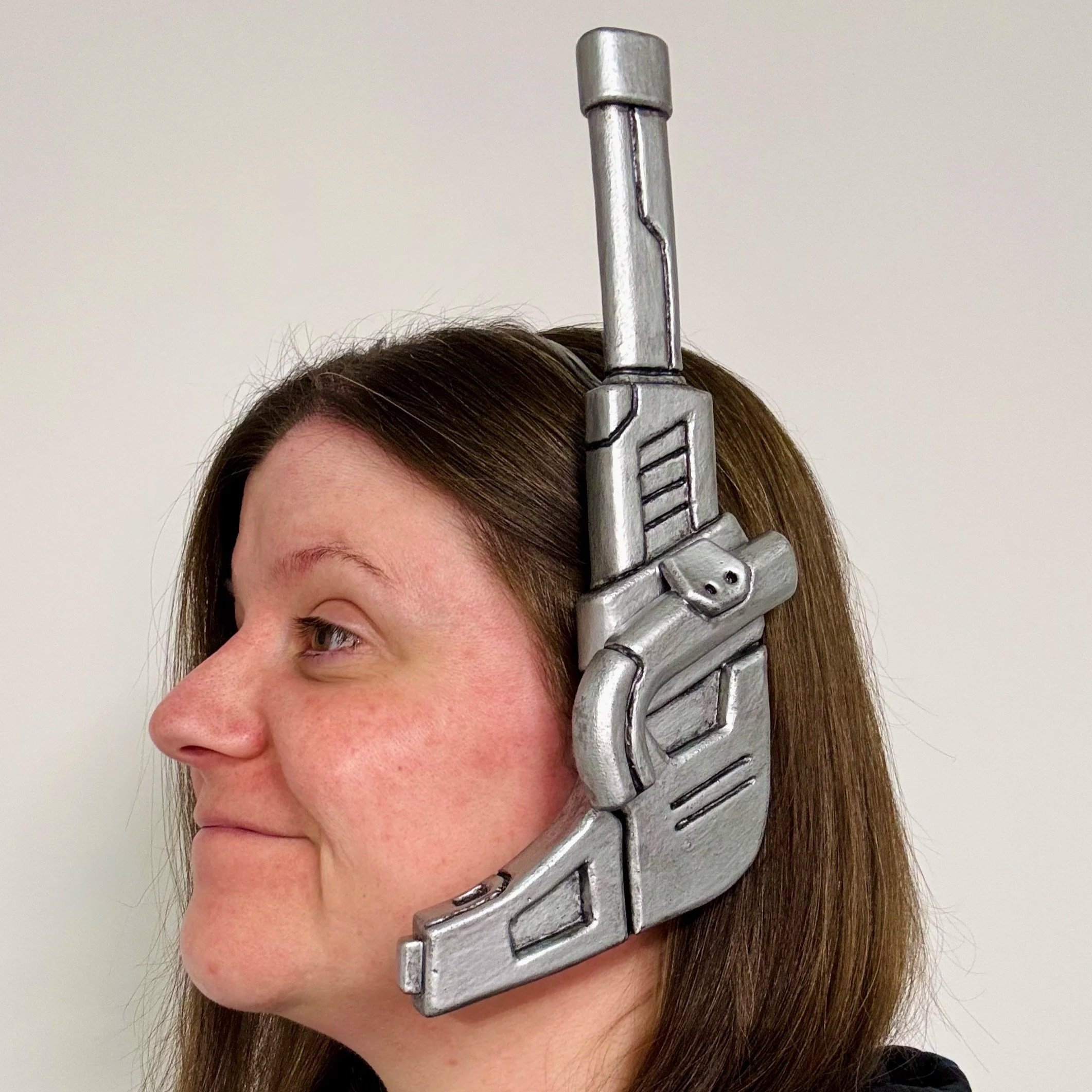 Close-up of a woman's face with a metallic superhero earpiece on her head against a plain white background.