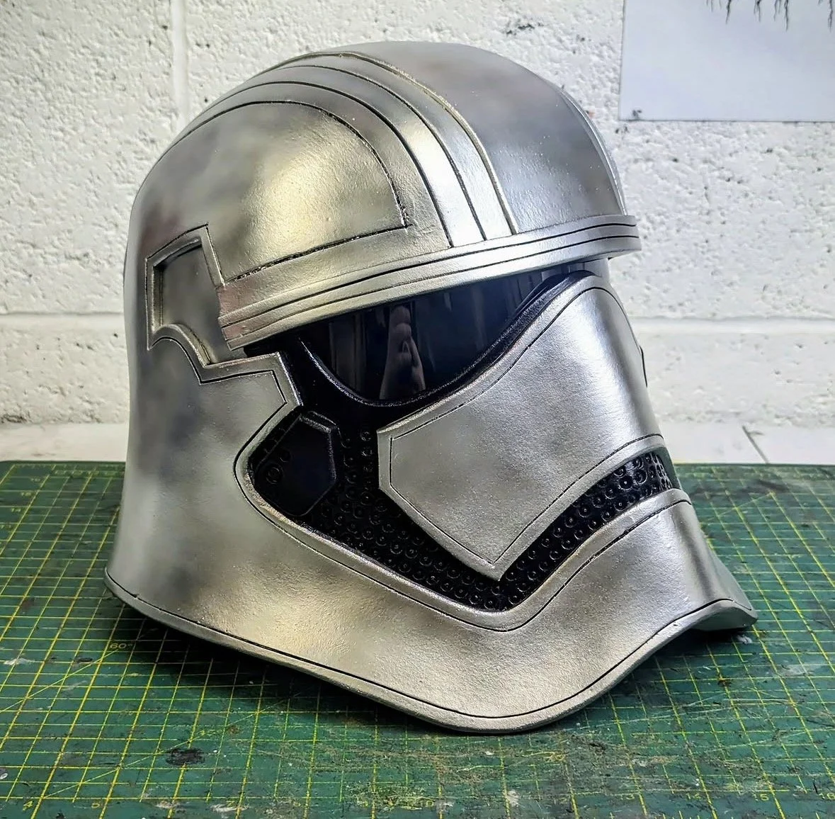 A silver, metallic helmet with a black visor, placed on a green cutting mat in front of a white brick wall.