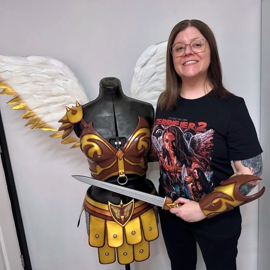 A woman standing next to a costume armor with large white angel wings, holding a sword, wearing a 'Terrifier 2' themed T-shirt.