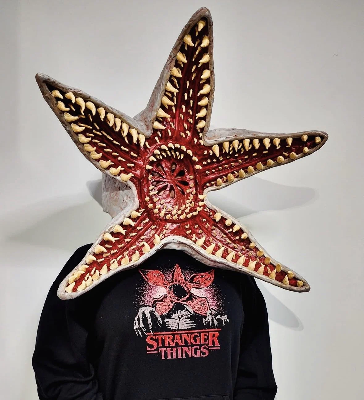 A person wearing a black hoodie with a 'Stranger Things' logo and graphic, with a large demogorgon mask that has red, white, and yellow details covering their face and head.