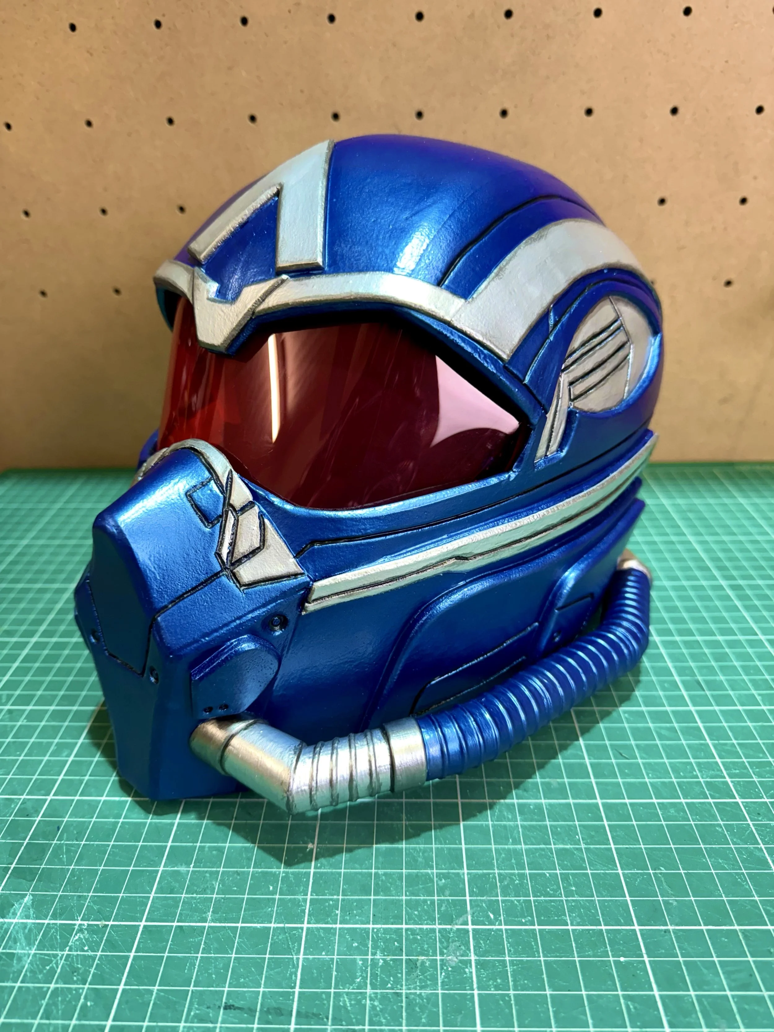 A superhero flight helmet helmet painted in blue with silver accents, featuring a red-tinted visor, displayed on a green cutting mat against a pegboard background.