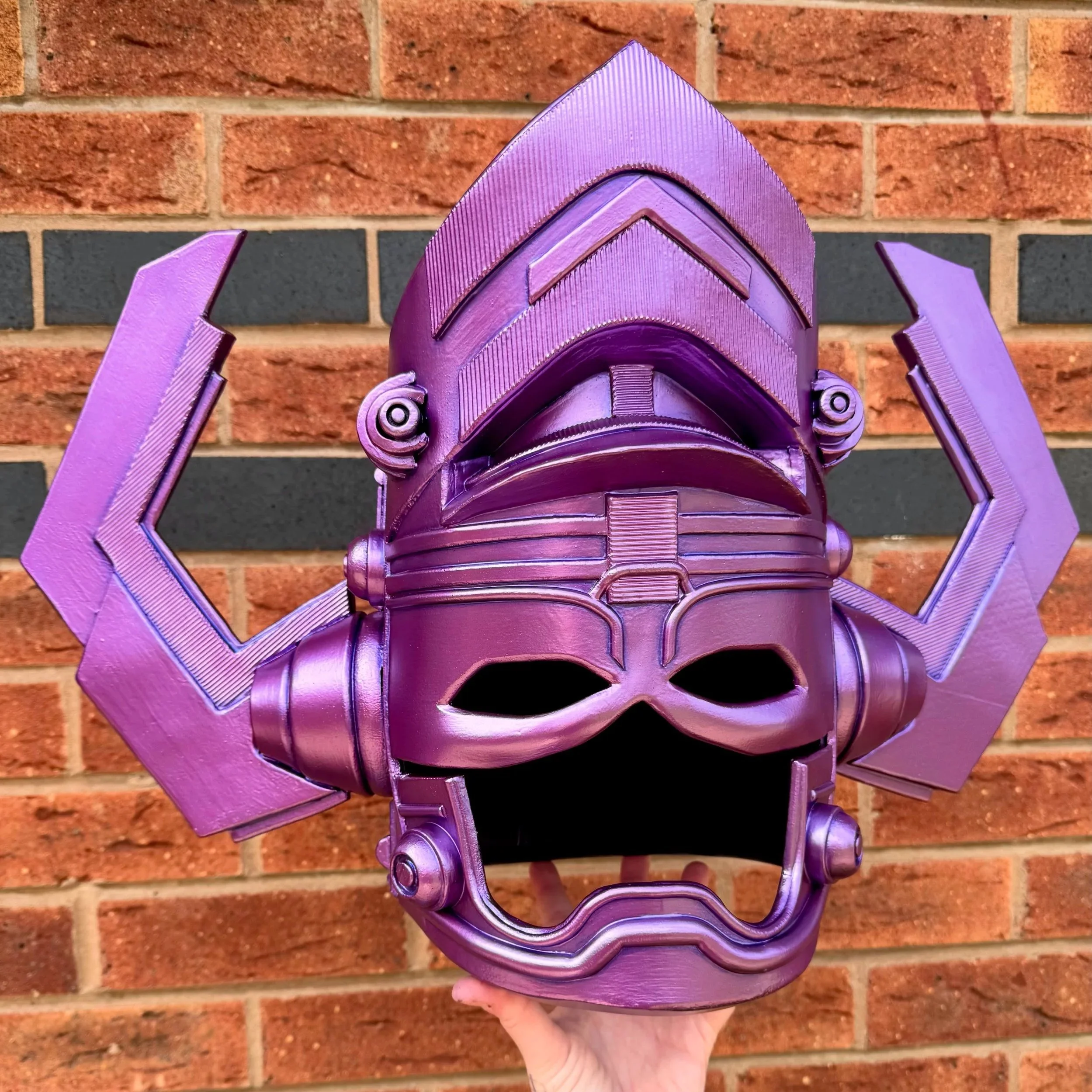 A person holding a purple, robotic-looking helmet against a brick wall background. The sculpture has geometric shapes and detailed textures, resembling Galactus from the Fantastic 4