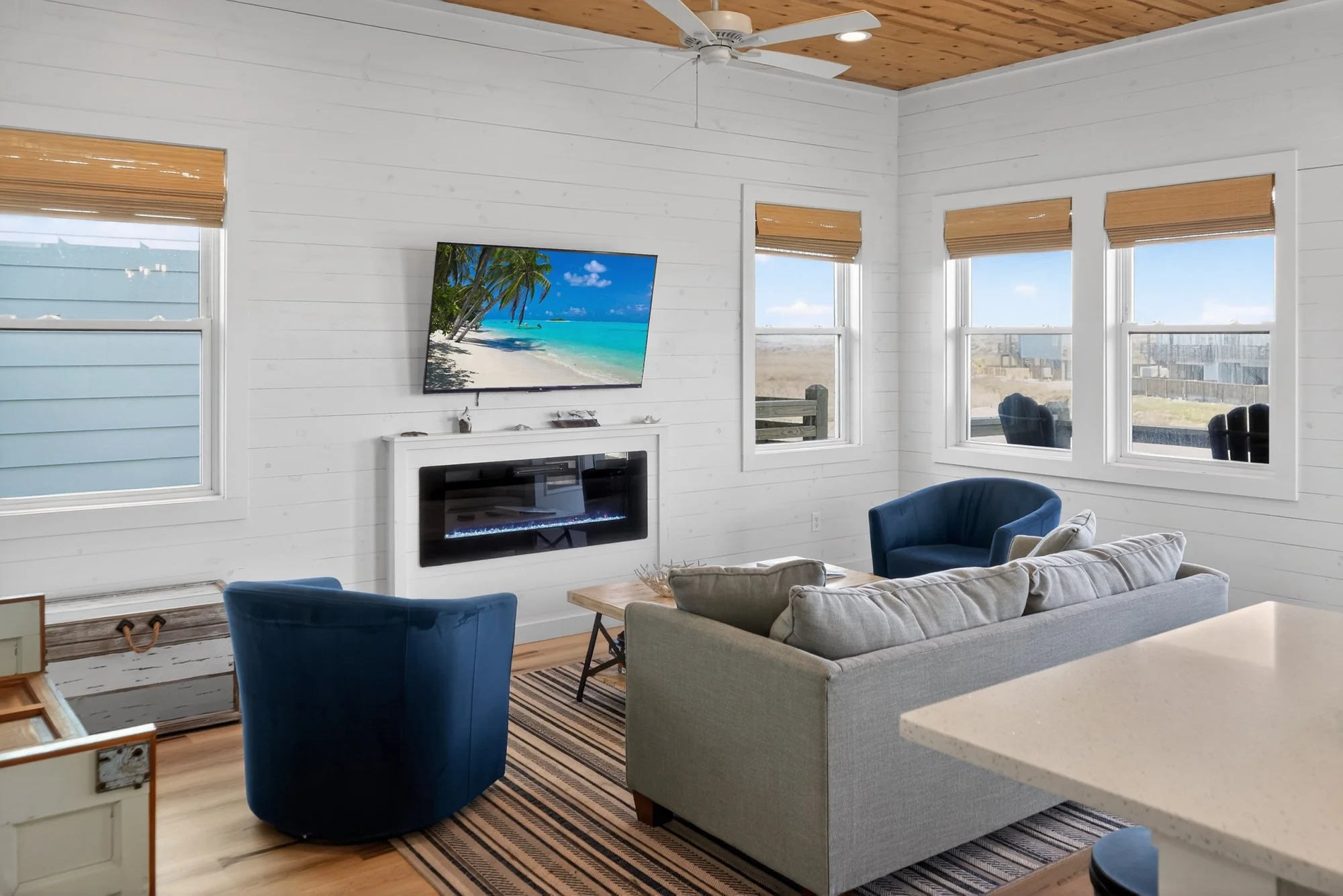 Living room with white walls, a flat-screen TV displaying a beach scene, a white fireplace, blue and beige chairs, a patterned area rug, large windows with wooden blinds, and a ceiling fan.