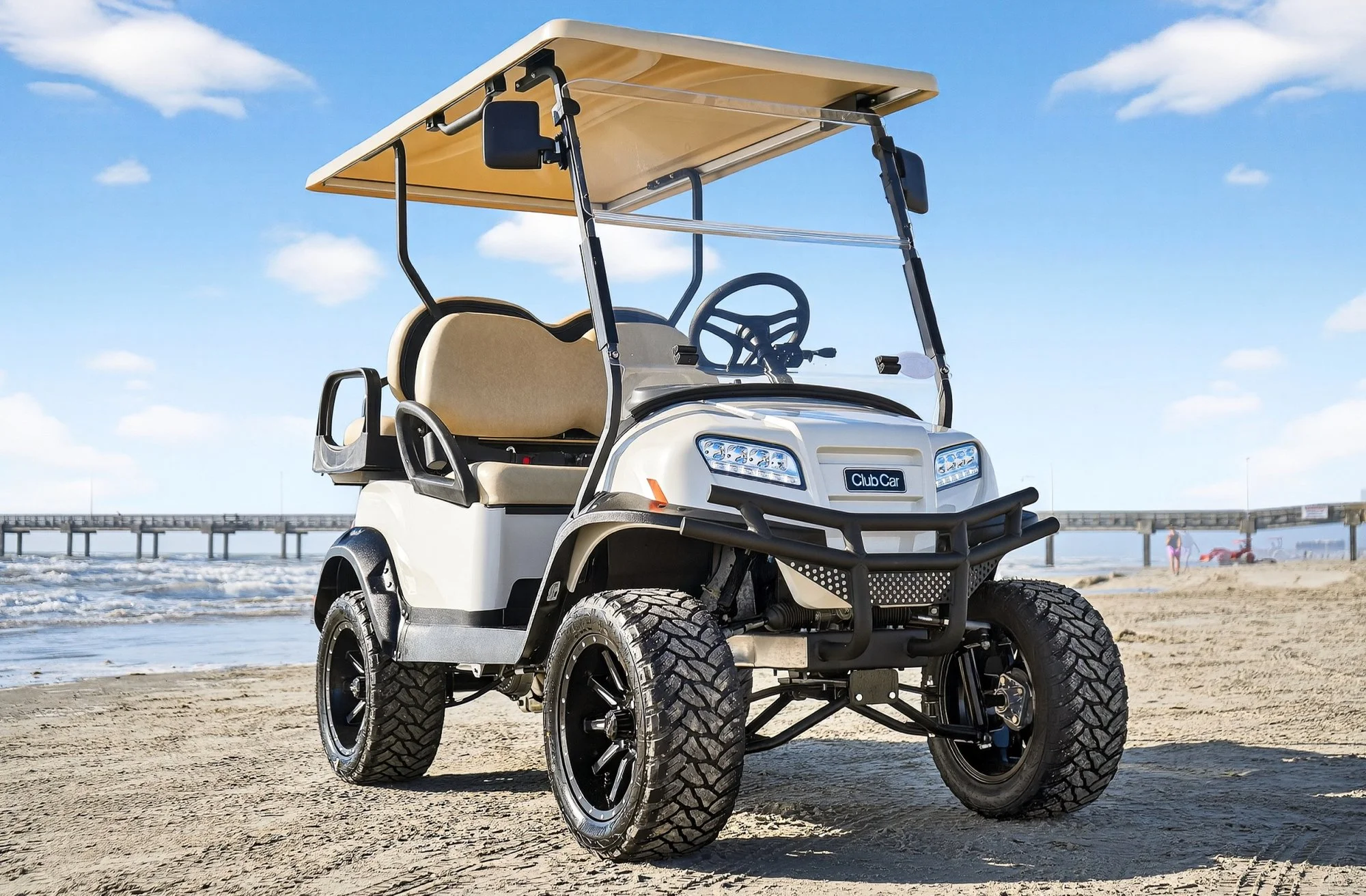A beige and black golf cart with large off-road tires parked on a sandy beach, with a pier and people in the background under a partly cloudy blue sky.