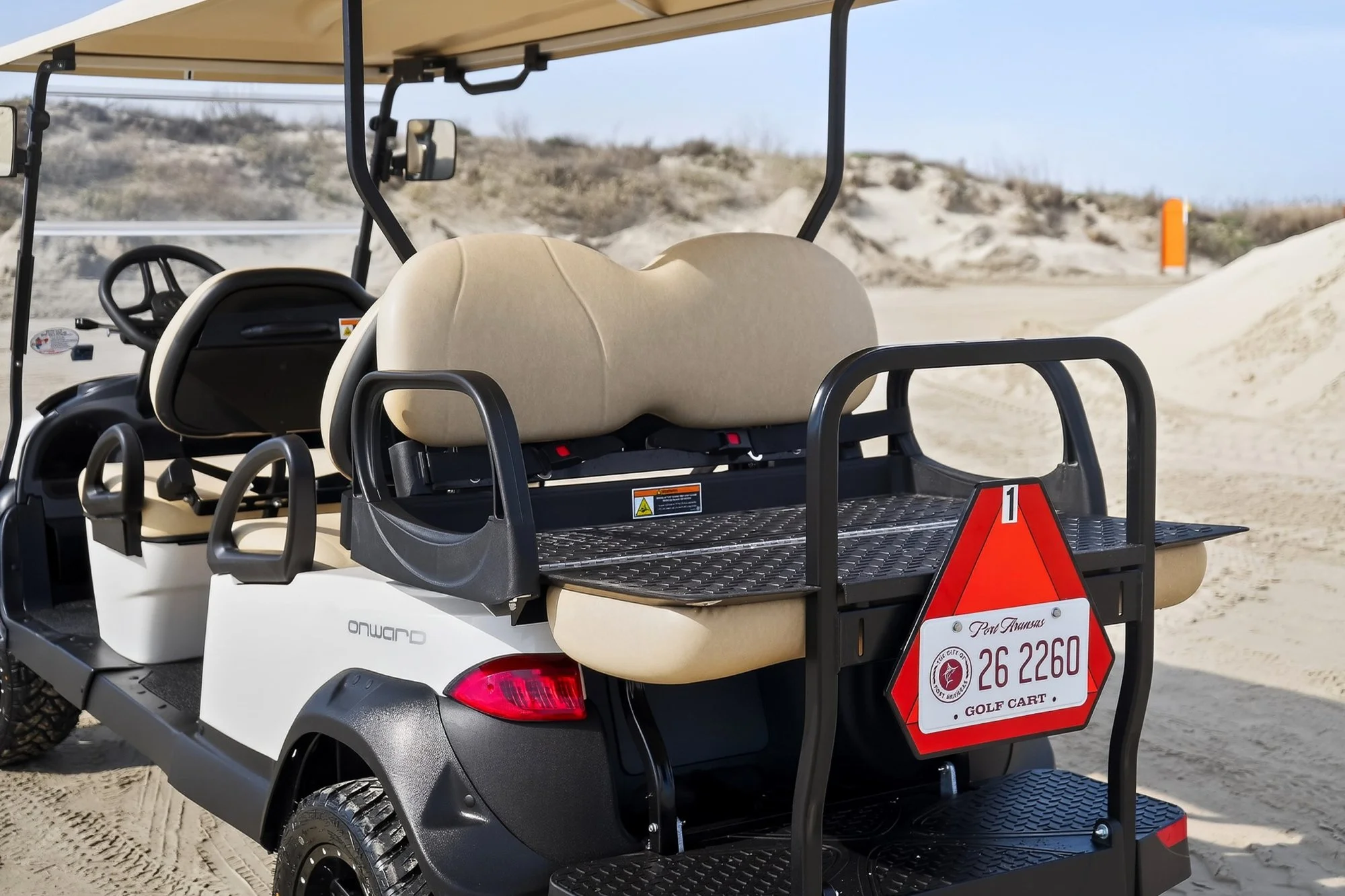 Golf cart parked on sandy terrain with a beige roof and black frame. It has two beige seats, a steering wheel, and a rear storage area with a warning sign. In the background, there are sand dunes and sparse vegetation under a clear blue sky.