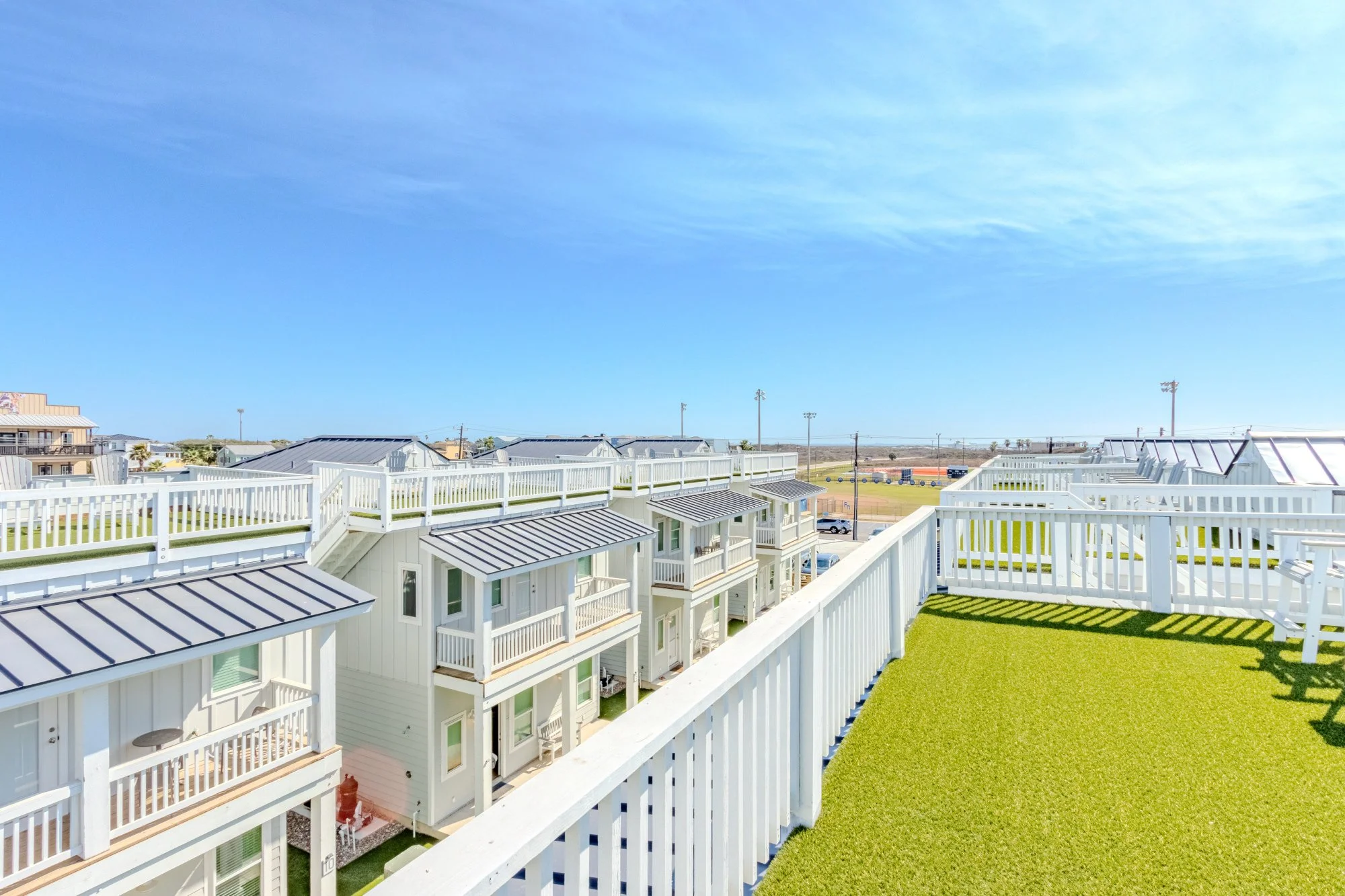 View from a balcony showing white railings, rooftops of white houses with small balconies, a green artificial turf on the balcony floor, and a distant sports field with orange ground and light poles under a blue sky with some clouds.