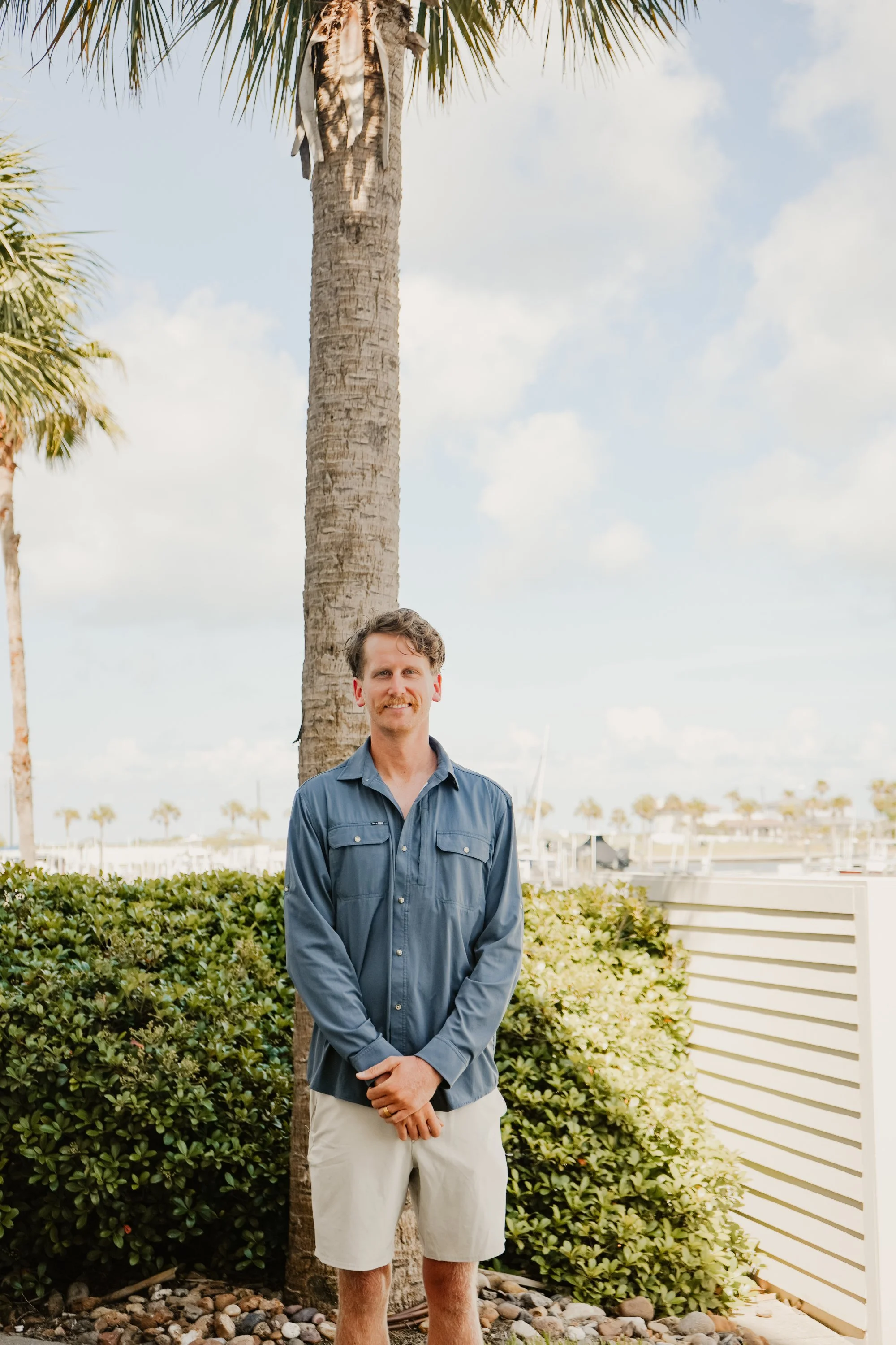 A man with light brown hair wearing a blue button-up shirt and beige shorts standing in front of a palm tree with a backdrop of cloudy sky and marina with boats.
