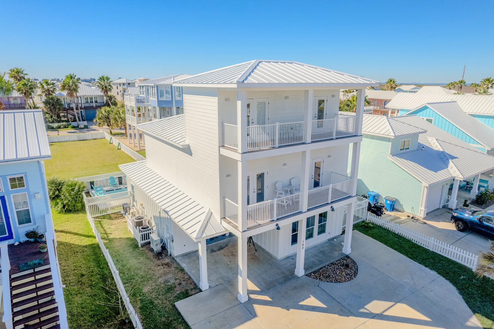 White multi-story beach house with balconies, metal roof, and outdoor patio area; neighboring colorful houses and palm trees in a coastal neighborhood under blue sky.