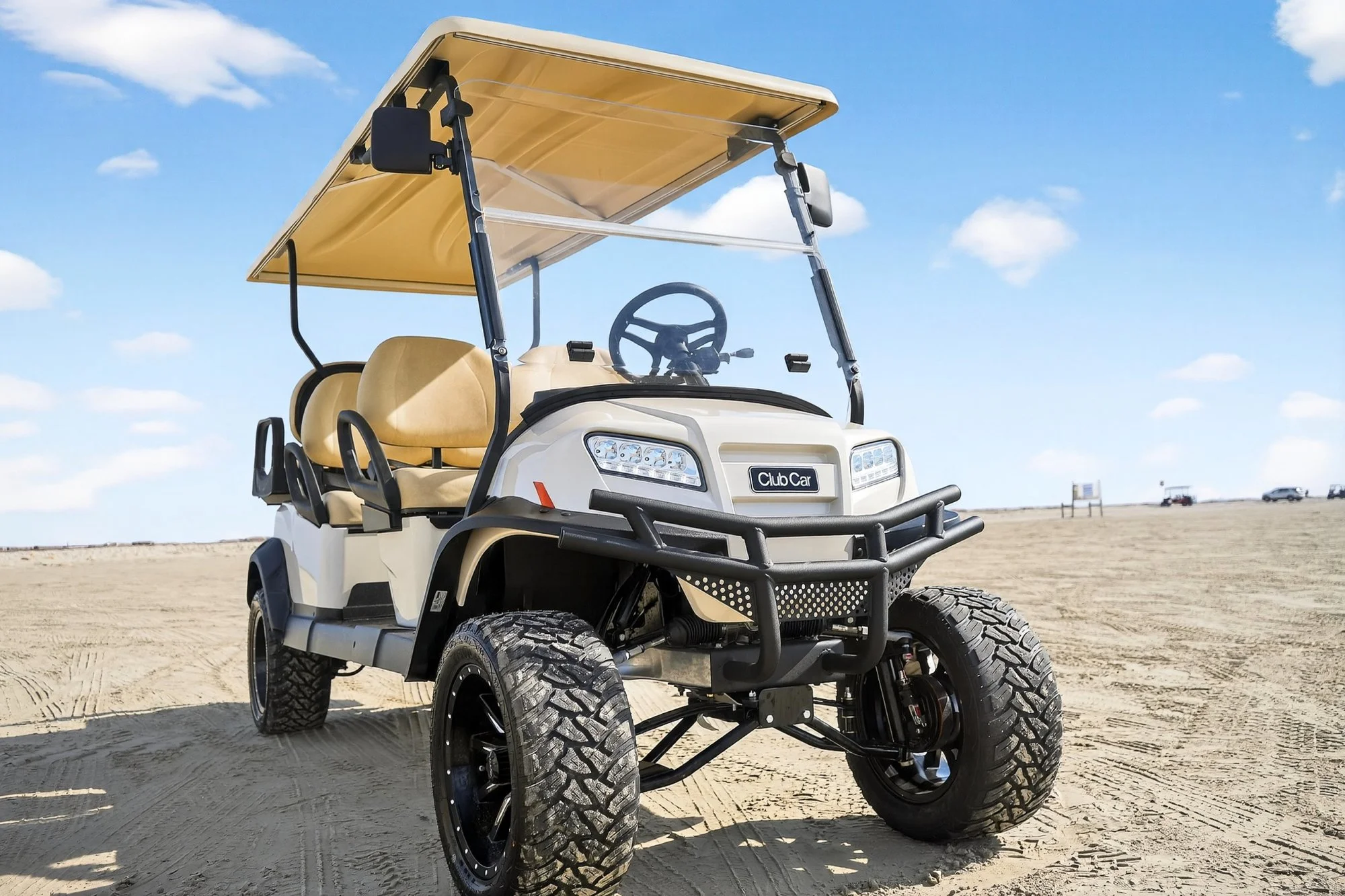 A beige golf cart with large off-road tires parked on sandy terrain under a blue sky with clouds.