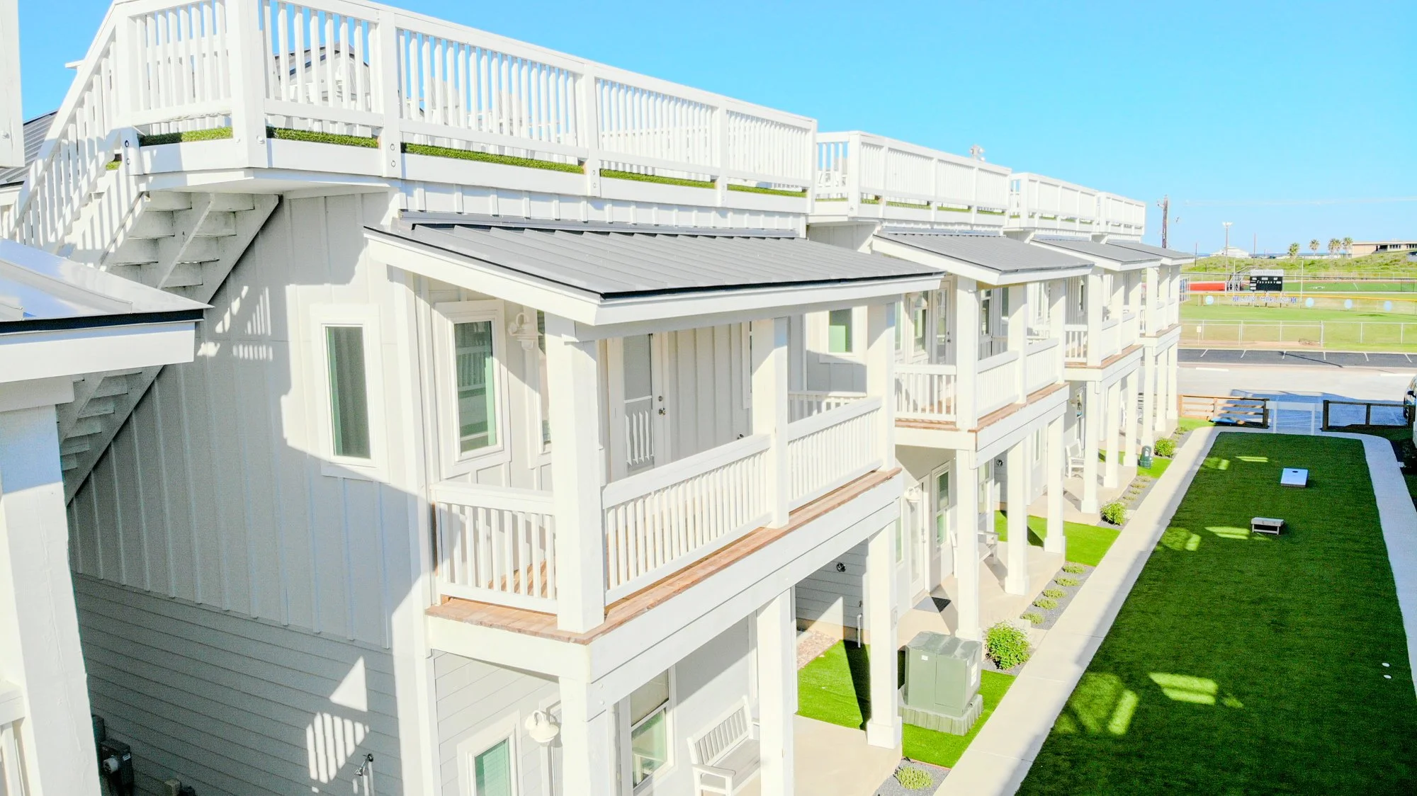White beachfront apartment building with multiple balconies and a rooftop deck, overlooking a grassy outdoor area and a baseball field, under a clear blue sky.