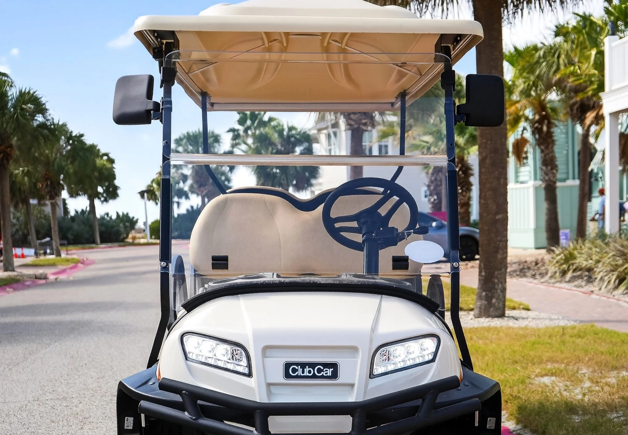 Front view of a white golf cart with a beige roof, black steering wheel, and a black bumper, parked on a street lined with palm trees and colorful buildings in the background.