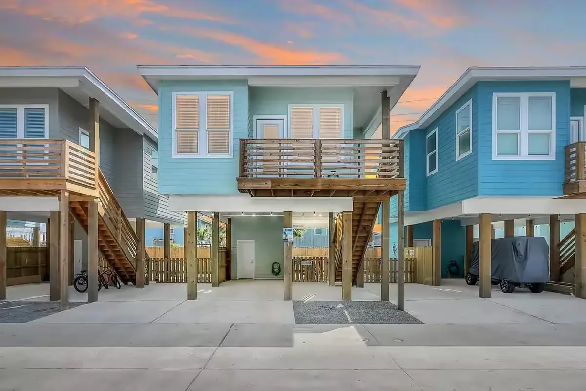 Three modern stilt houses in pastel blue shades with wooden stairs and balconies, set against a colorful sunset sky, with parking spaces below and bikes and covered vehicles.