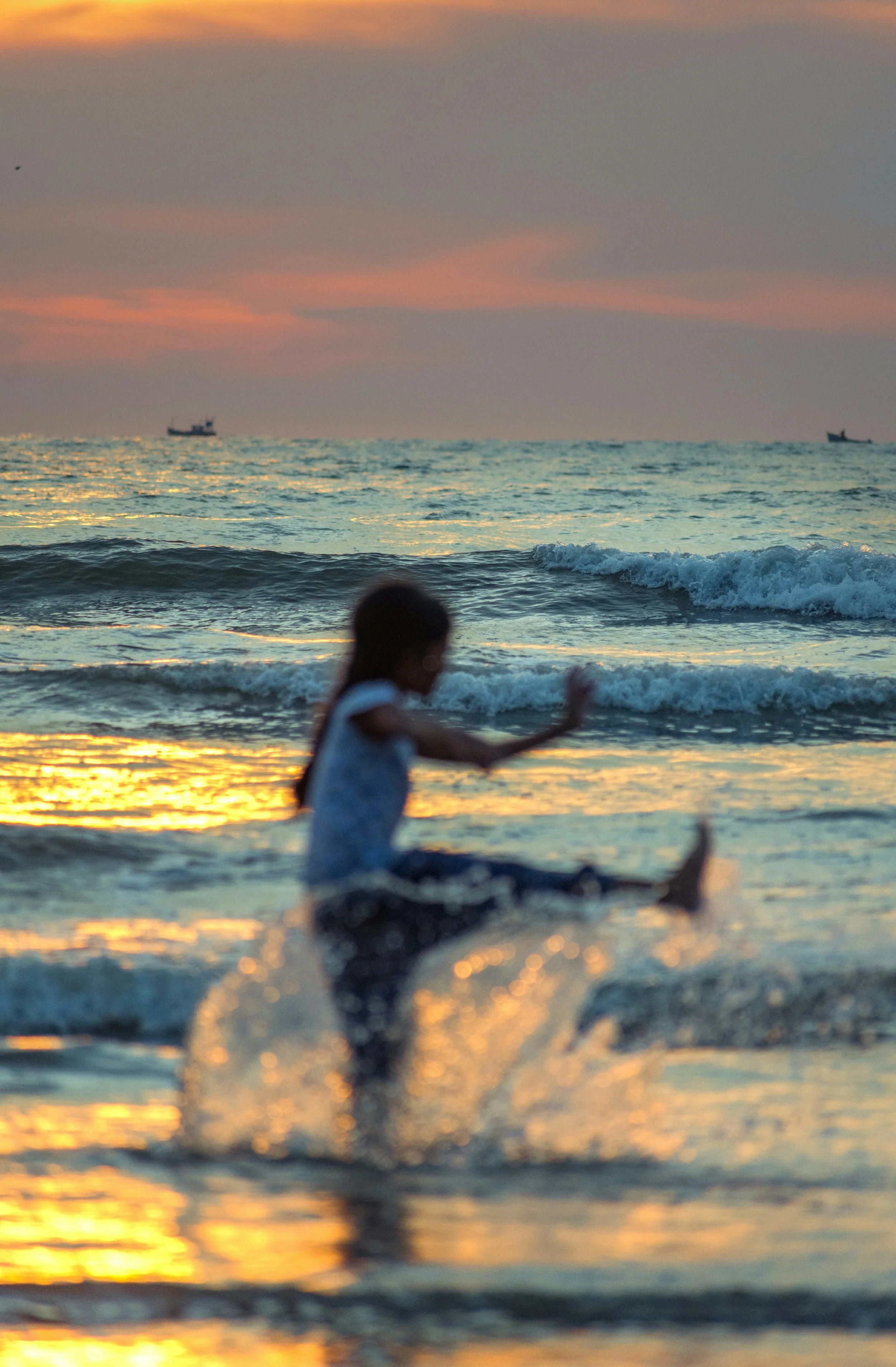Child playing in the ocean during sunset with ships on the horizon.