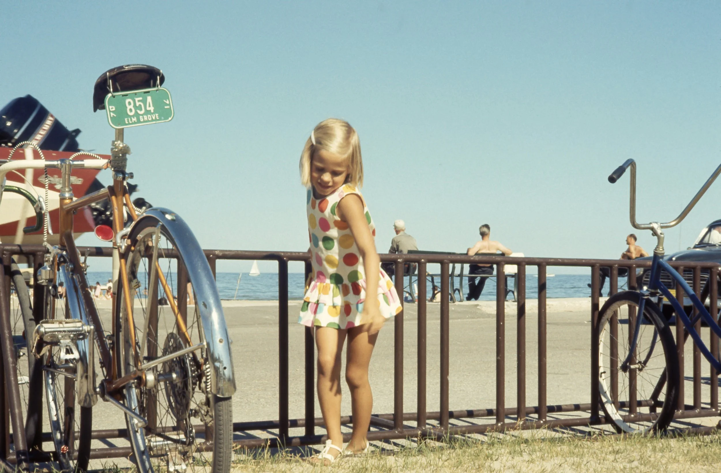 A young girl in a colorful polka dot dress walking near a railing at the beach on a sunny day, with bicycles parked nearby and people in the background by the water.