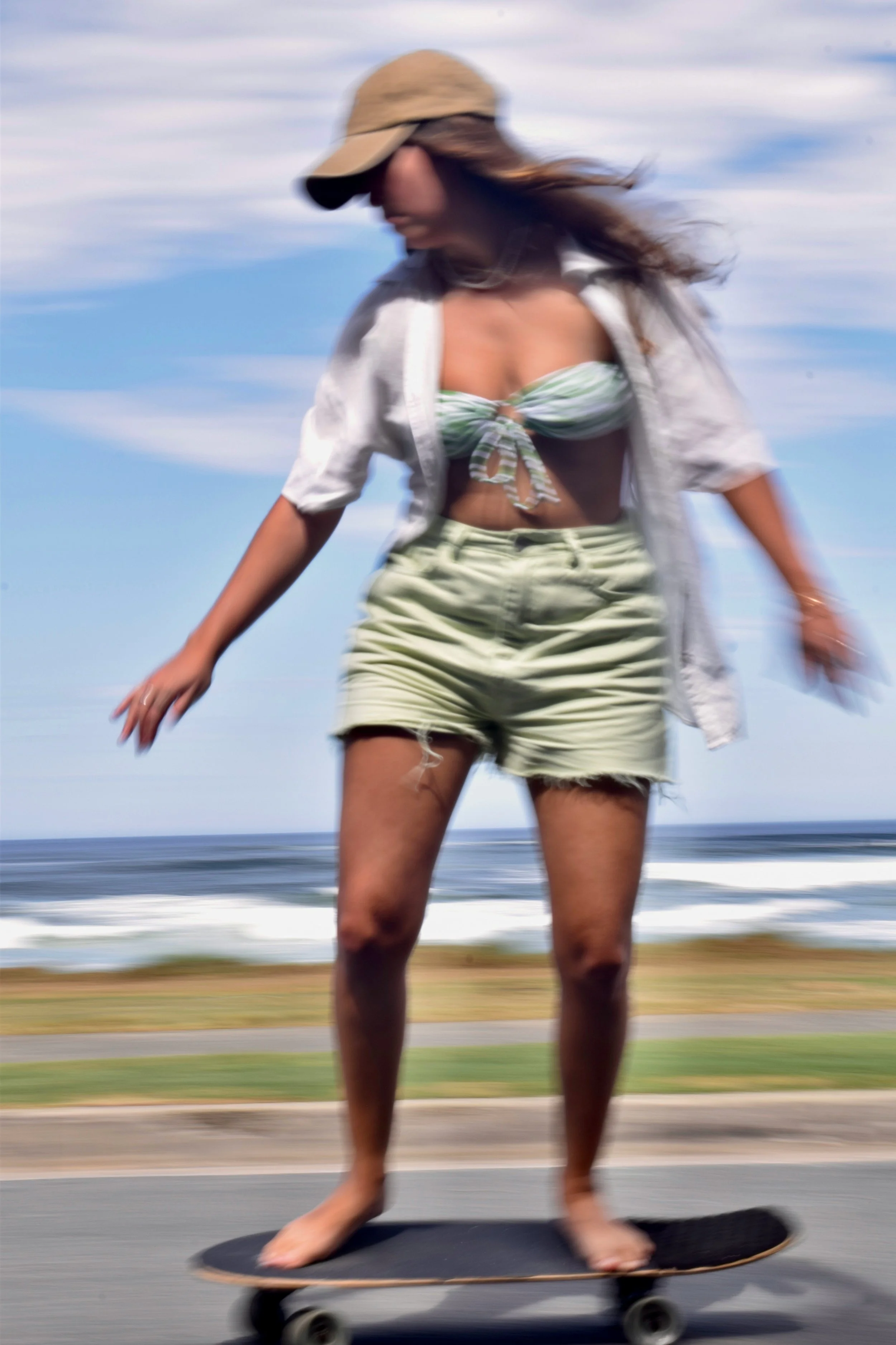 A woman skateboarding on a beachside road, wearing a beige hat, white shirt, green striped bikini top, and light-colored shorts, with ocean waves in the background and a partly cloudy sky overhead.
