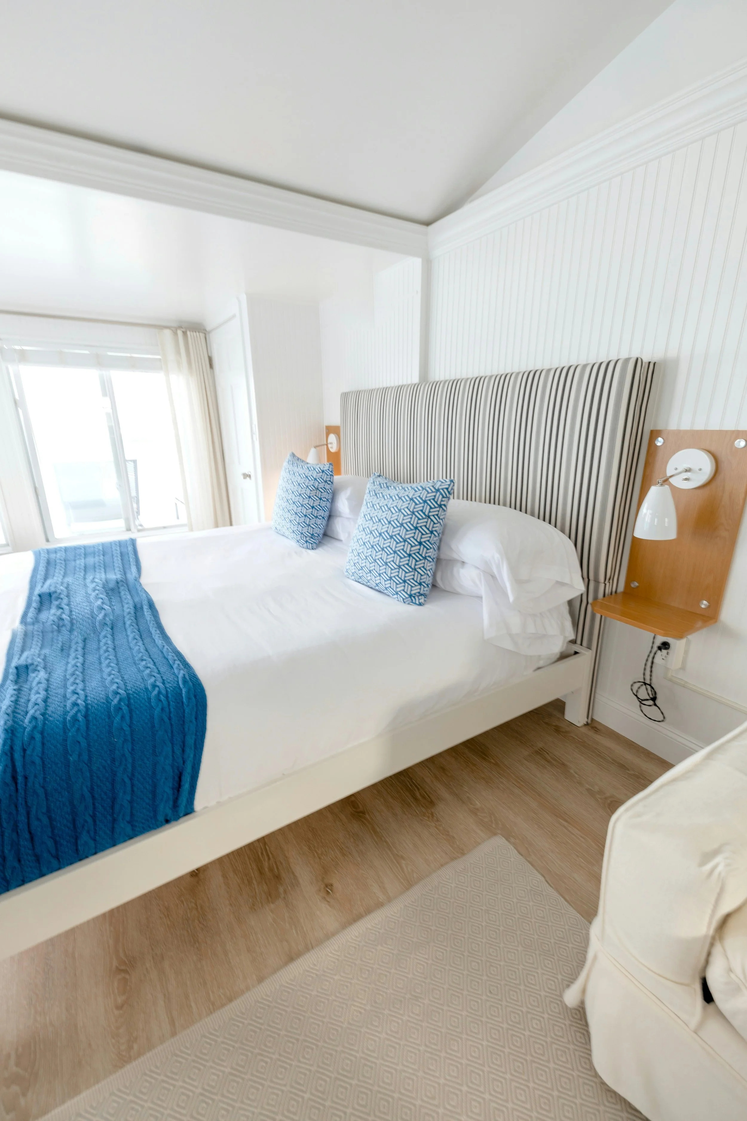 A bedroom with a bed, blue blanket, and two patterned blue pillows. The headboard is striped fabric, and there are wall-mounted lamps on either side. The room has light wood flooring and a beige textured rug.