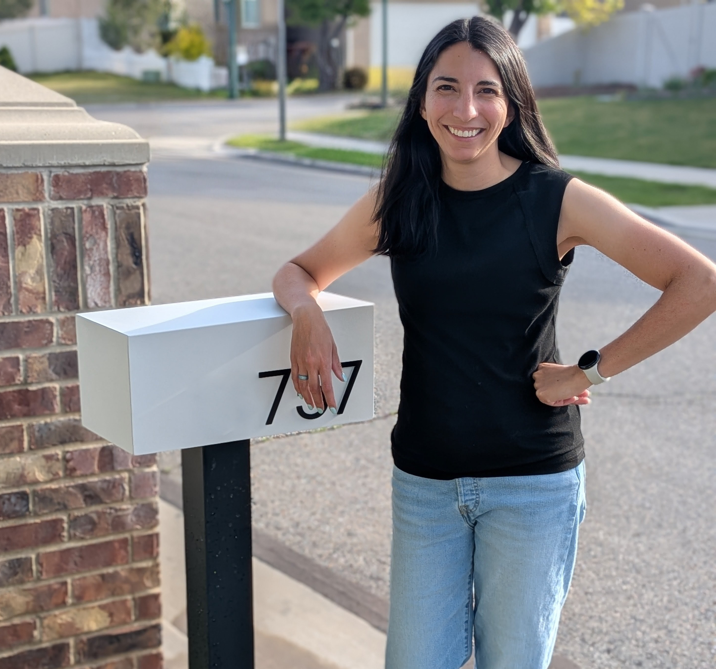 A woman with long dark hair, wearing a sleeveless black top and blue jeans, standing outdoors on a sidewalk next to a white post with the number 753. The background features a residential neighborhood with trees, a street, and houses, during daylight.