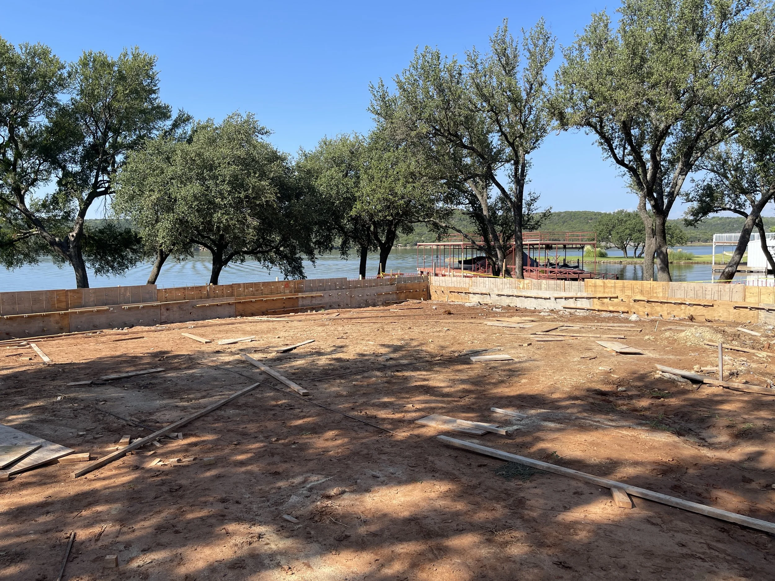Construction site by a lake with trees and a clear blue sky, showing wooden framing on the ground and partially built structures in the background.