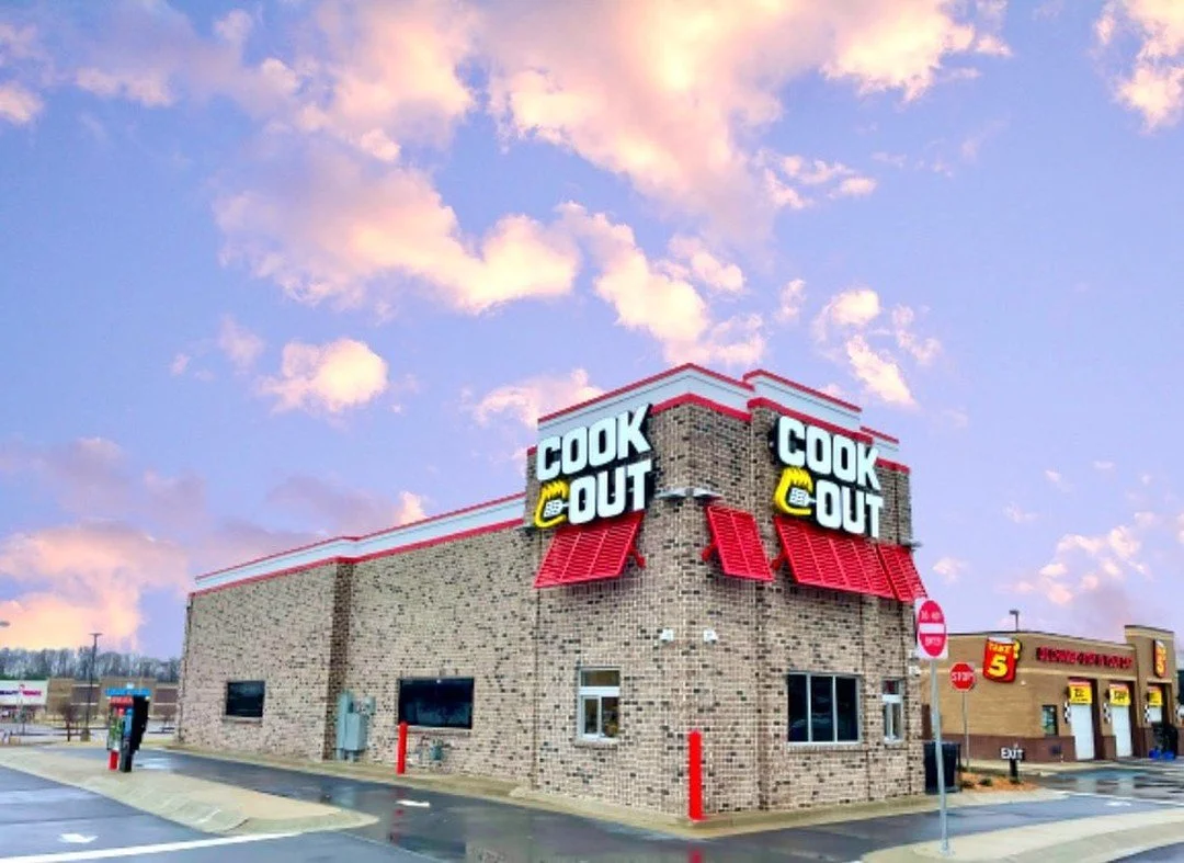 A fast-food restaurant named Cook Out with brick exterior, red awnings, and illuminated signs on the building, under a sky with pink and orange clouds.