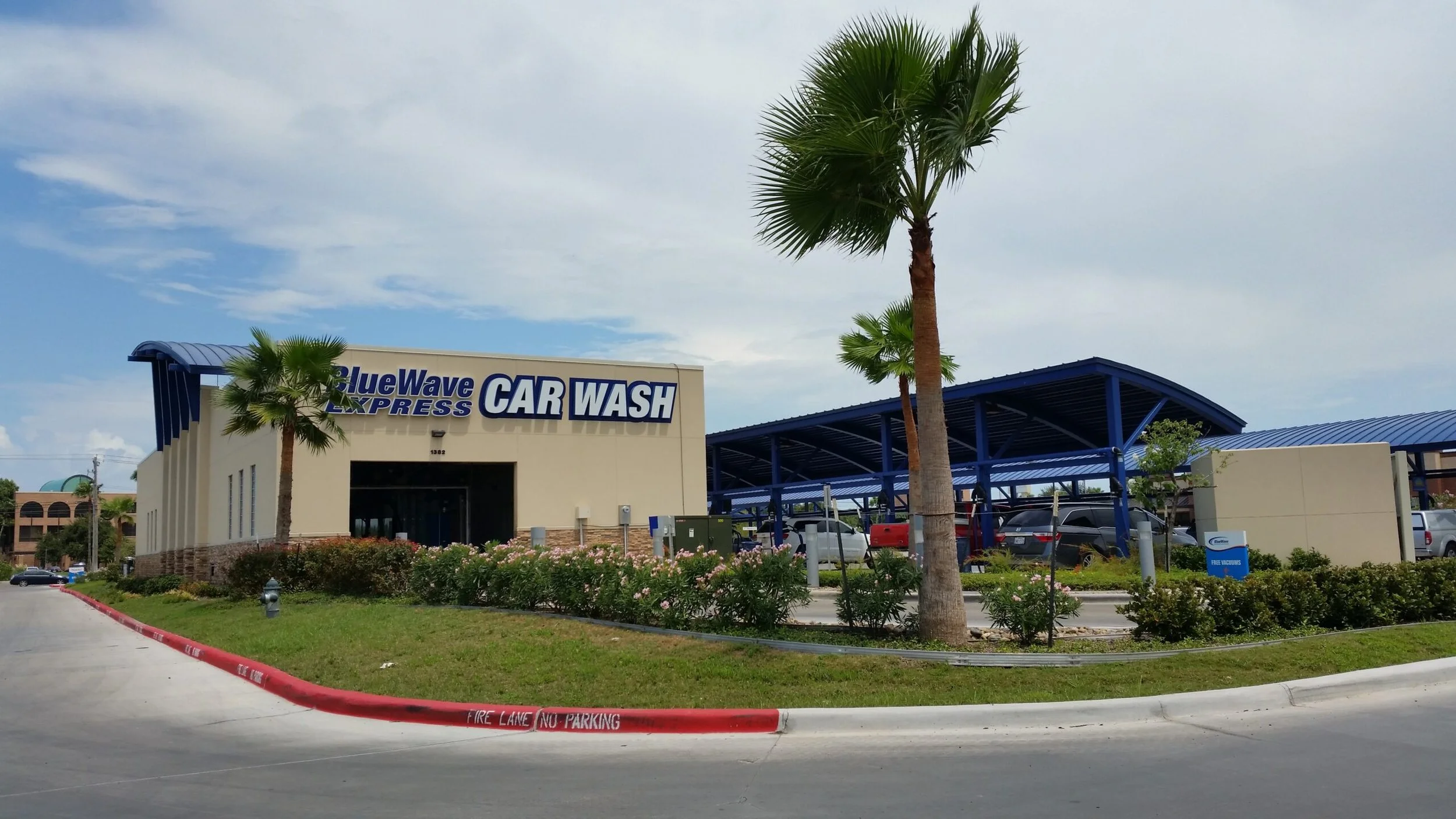 Exterior of BlueWave Express Car Wash building with palm trees in front, parked cars, and a blue canopy, under cloudy sky.