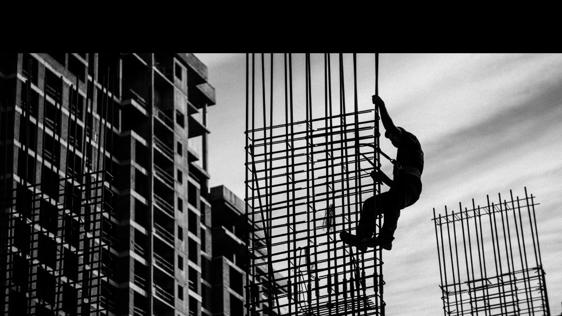 Black and white photo of a construction worker climbing rebar on a building site with a cityscape in the background.