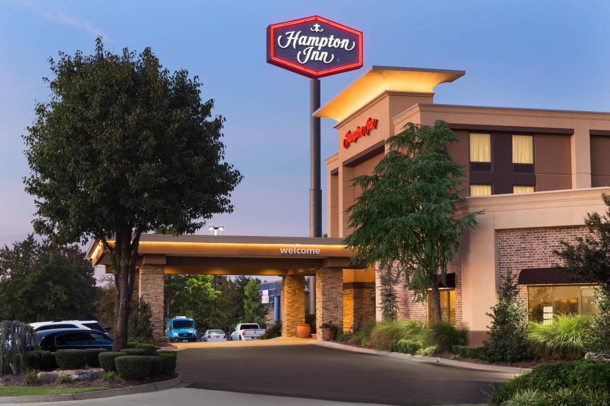 The entrance of a Hampton Inn hotel with a welcome sign and parking lot, surrounded by trees and landscaping, during dusk.