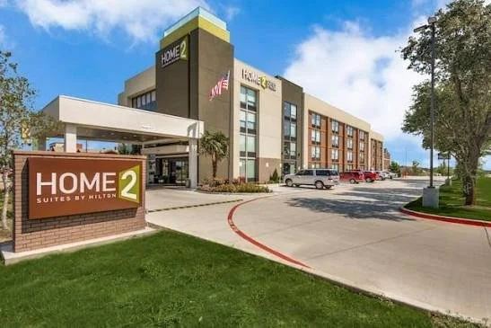 Exterior of a Home2 Suites hotel with a parking lot and some trees.