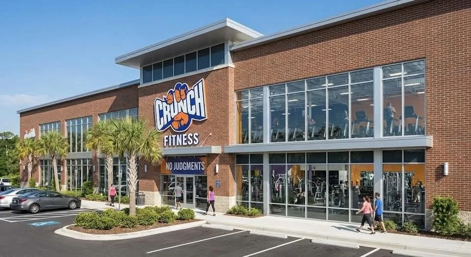 Exterior of Crunch Fitness gym with people walking and cars parked in front, large glass windows showing gym equipment inside, palm trees, and clear sunny sky