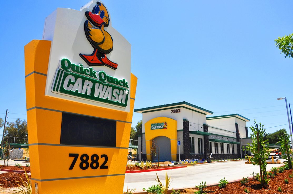 Exterior of a Quick Quack Car Wash with a colorful sign featuring a cartoon duck, yellow and white building, and clear blue sky.