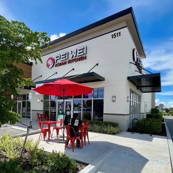 Exterior of a restaurant named PEI WEI Asian Kitchen with outdoor seating, red umbrella, and green landscaping under a blue sky.