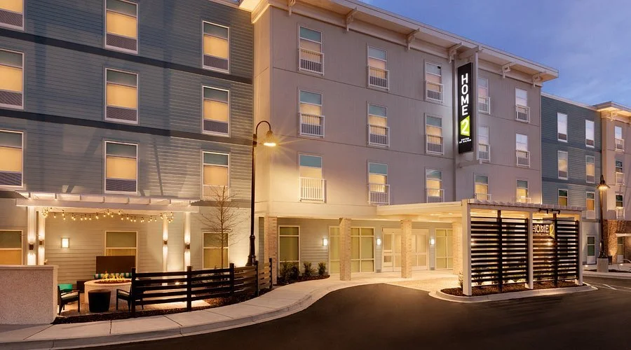 Exterior view of a modern hotel at dusk with a lit entrance, street lamps, and a sign reading 'Home2 Suites' on the building.