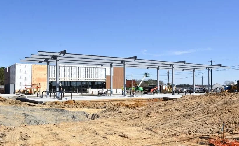 Under construction building with a steel frame and partially finished walls, with construction equipment and parked cars nearby.