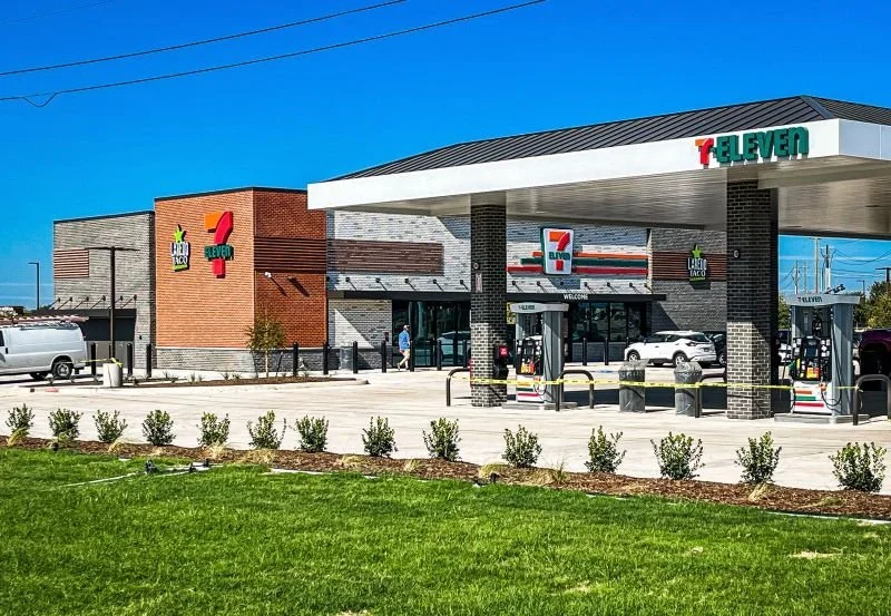 Exterior of a 7-Eleven convenience store with gas pumps in front, brick and modern siding, and some small shrubs in the foreground.