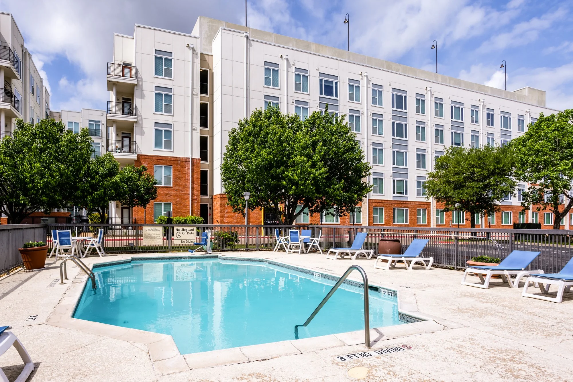 Swimming pool area with lounge chairs and tables, surrounded by a metal fence, in front of a modern multi-story apartment building with trees.