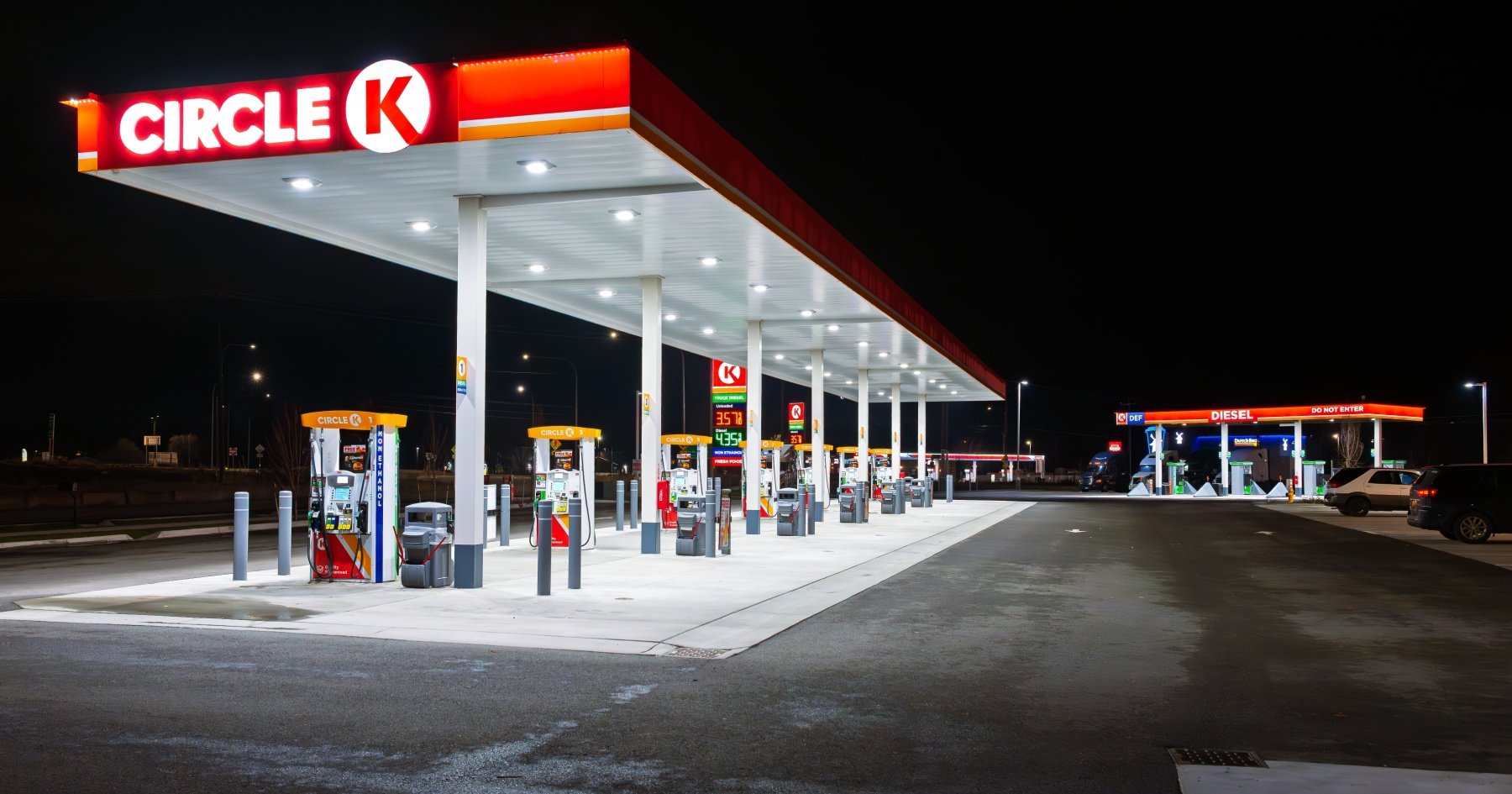 Night view of a Circle K gas station with multiple fuel pumps under a large red and white canopy, illuminated signs displaying fuel prices, and a separate diesel pump area with lit signs and parked cars.