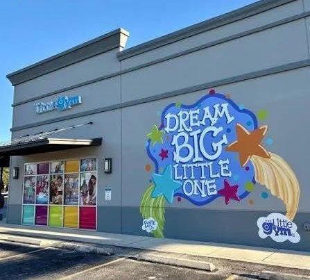 Exterior of a gym with colorful mural saying 'Dream Big Little One' and a window display of children and nutritional products.