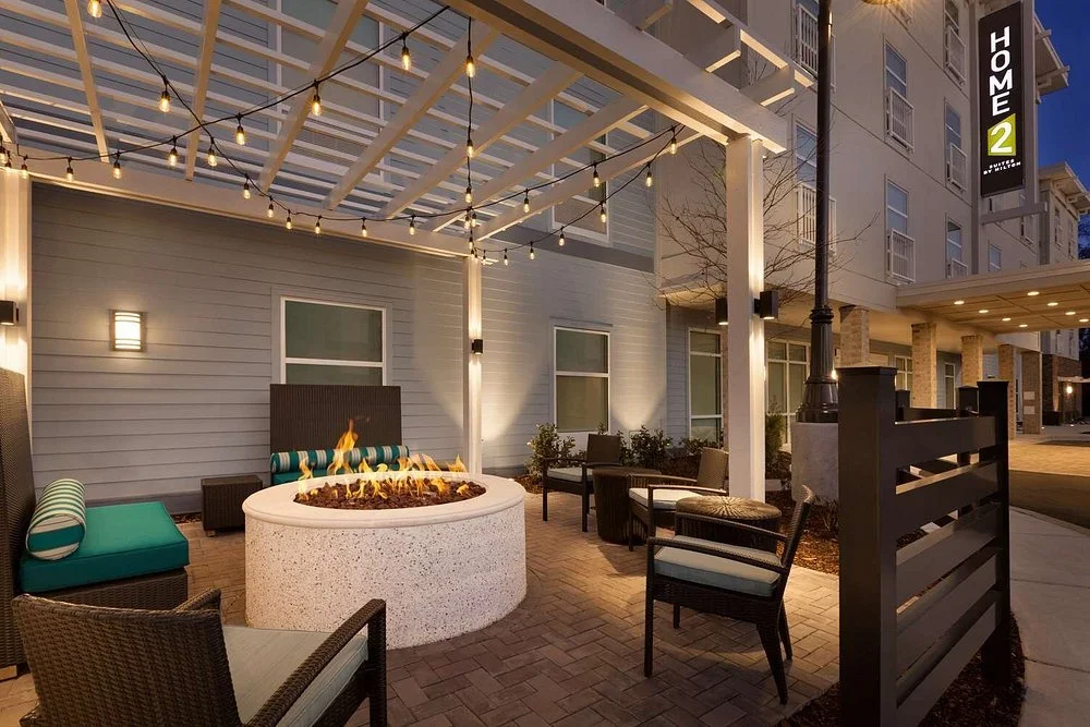 An outdoor patio area with a fire pit, string lights overhead, and chairs and tables around it, in front of a modern apartment building.