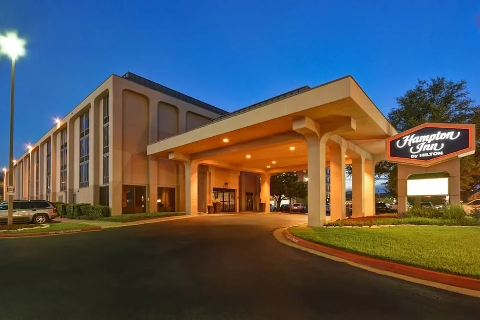 Exterior view of Hampton Inn hotel at dusk, showing the entrance, parking lot, and illuminated sign.