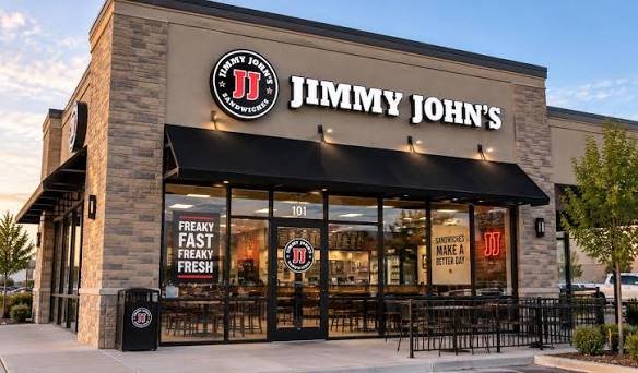 Exterior view of a Jimmy John's fast-food restaurant with a brick facade, black awning, and large glass windows, featuring the Jimmy John's logo and signage.