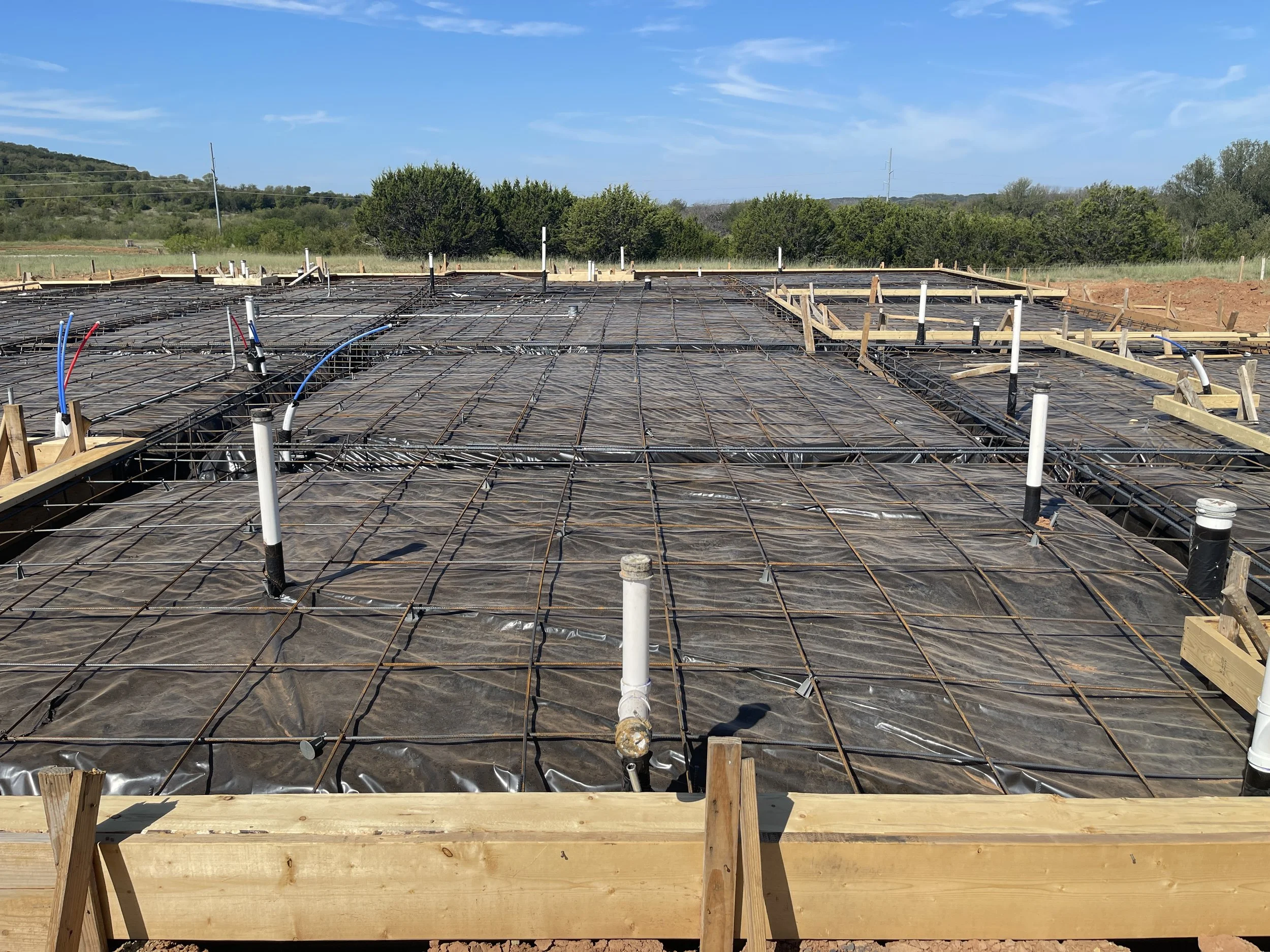 Construction site with foundation and plumbing pipes, surrounded by wooden beams and rebar, under a blue sky with some clouds.