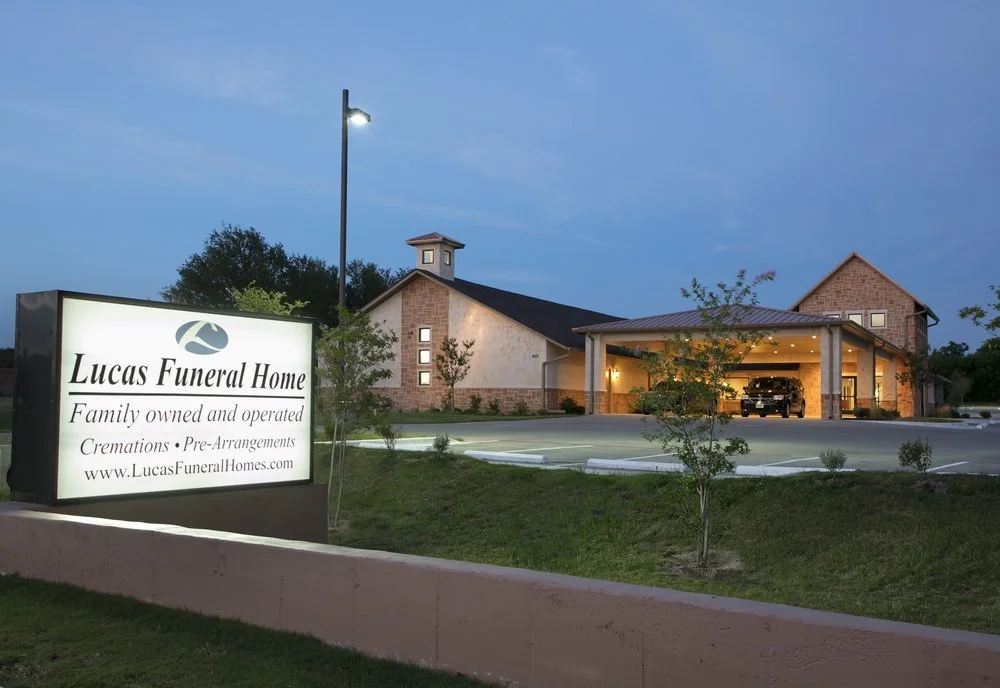 Exterior of Lucas Funeral Home building in the evening, with a lit sign displaying the funeral home's name and services, surrounded by a parking lot and small trees.