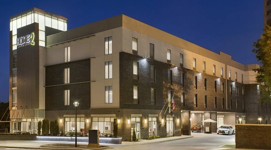 Exterior view of a modern mid-rise building at dusk with lit windows, street lamps, and a parking area with cars, featuring a hotel sign that reads 'Home2 Suites'.