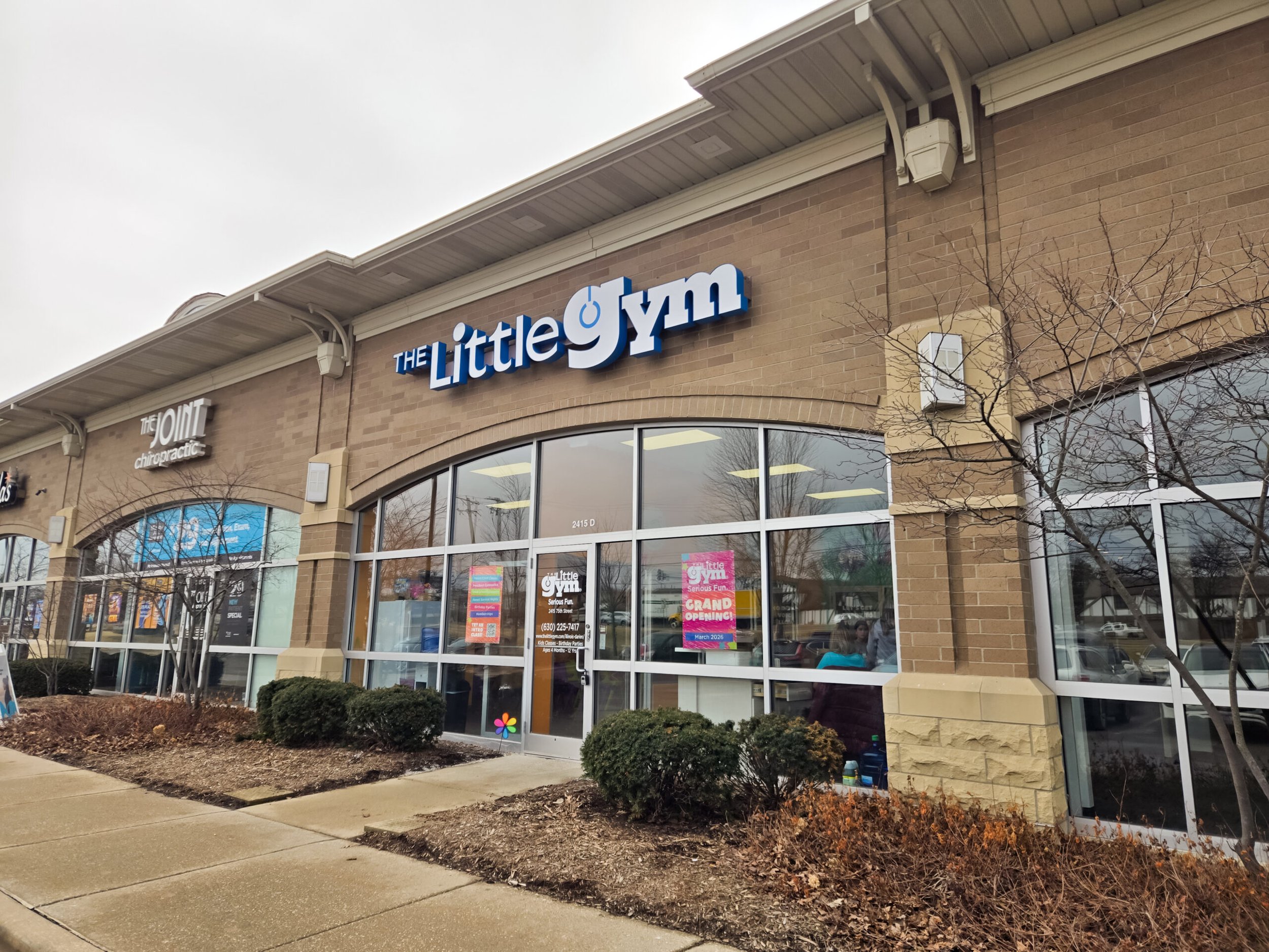 The storefront of The Little Gym, a children's fitness center, with large windows displaying promotional signs and banners advertising grand opening events. The building has brick walls and a sidewalk with small bushes and bare trees in front.