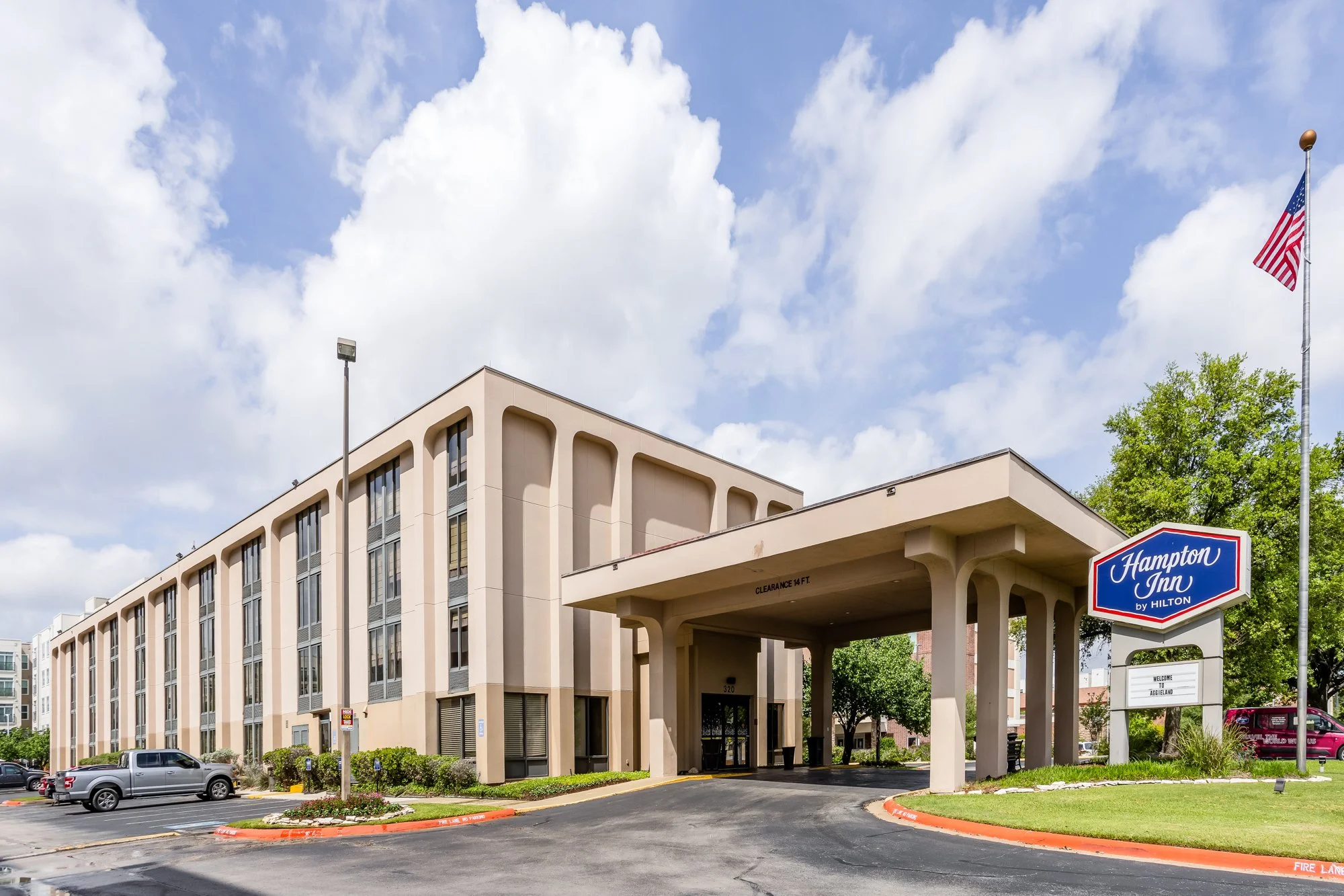 Exterior view of the Hampton Inn by Hilton hotel with a parking lot in front, American flag on a flagpole, trees, and a partly cloudy sky.